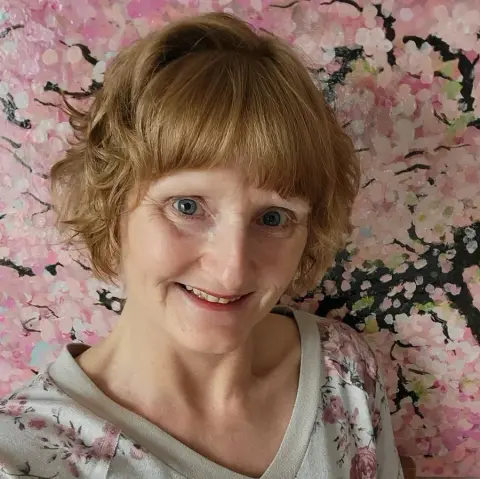 A woman with short curly red hair and blue eyes smiling in front of a background with pink cherry blossom artwork.