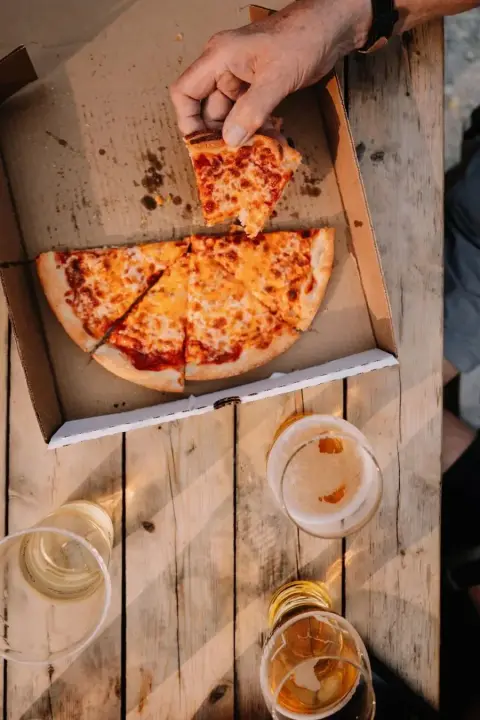 A hand is removing a slice of cheese pizza from an open pizza box on a rustic wooden table. There are three glasses of beer on the table, so