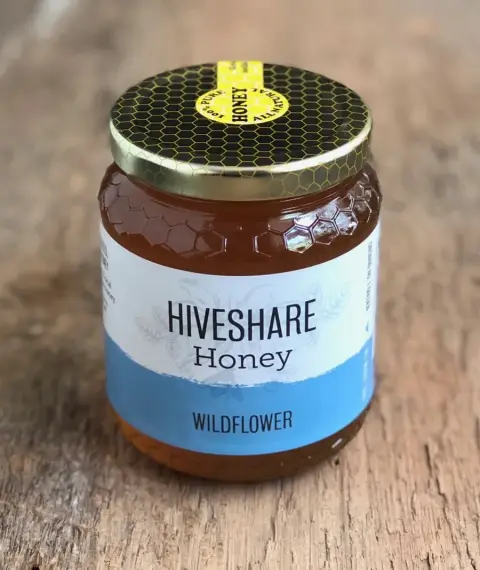 Image contains a glass jar of wildflower honey, with a white and blue rectangular label.