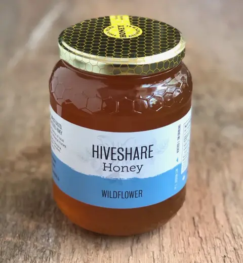 Image contains a large, 1kg glass jar of wildflowerhoney, on a wooden surface. The honey is a golden yellow colour, and the label is white a