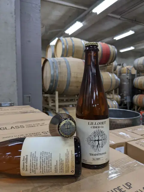 A bottle of Lillooet Cider Co. Traditional cider in front of oak barrels