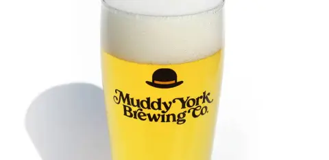 A glass of golden lager with the Muddy York bowler-hat logo