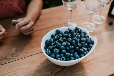 A bowl of fresh blueberries on a wooden table with three empty wine glasses in the background and a person holding a blueberry in their hand