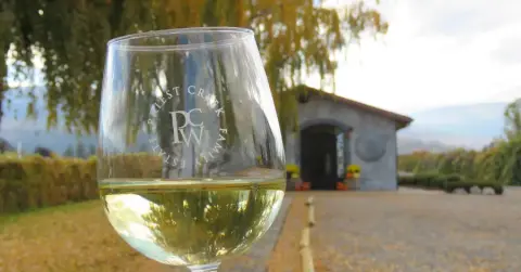 A glass of Priest Creek white in front of the stone tasting room in autumn