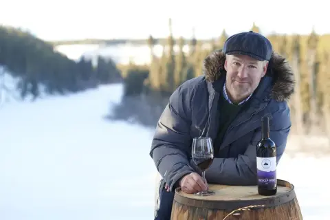 Founder Kyle Marchuk with a bottle and glass of Solstice haskap wine on a barrel above a snowy Yukon river valley