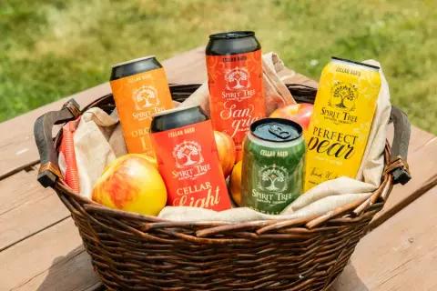 A basket of Spirit Tree cider cans, Cellar Light, Crabapple Sangria, Perfect Pear and Premium, with fresh apples