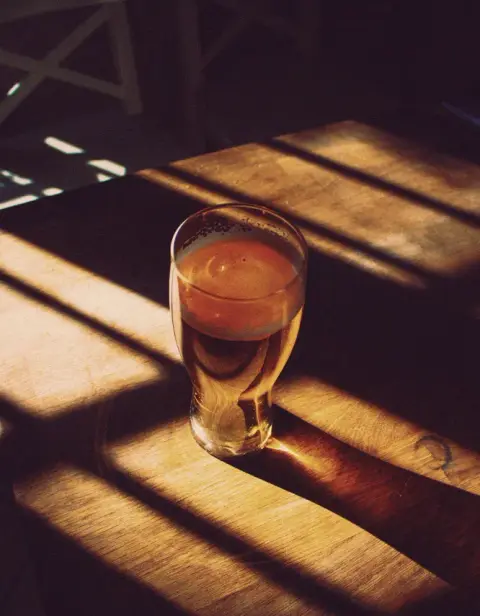 A pint glass with a blonde ale sitting in the sun on a wooden table