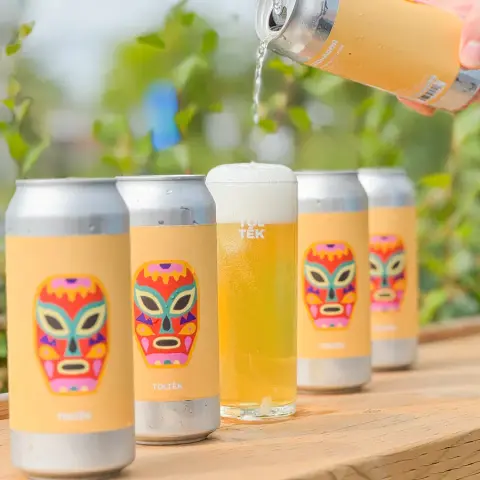 Toltèk cans with a colourful mask label beside a freshly poured glass of beer