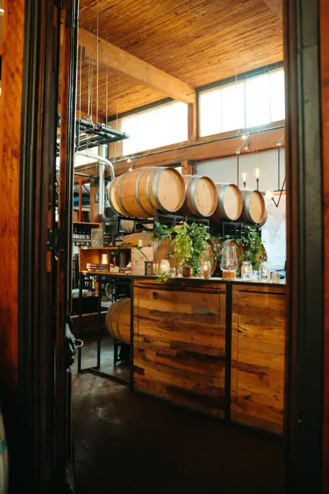Inside a rustic winery or tasting room with wooden barrels stacked on shelves and a wooden counter decorated with plants and candles, illumi