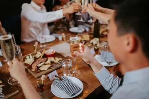 People clinking glasses at a dinner party with a charcuterie board, cheese, and drinks on a wooden table.