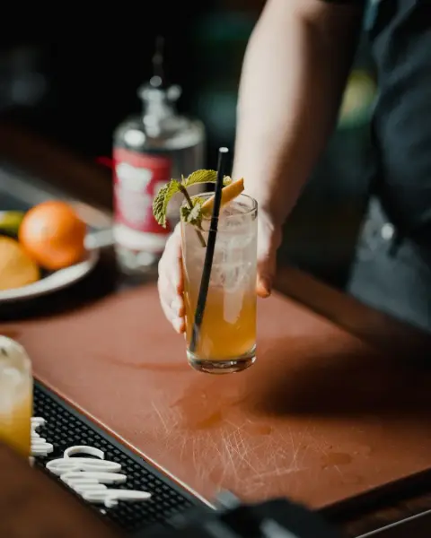 Bartender holding a cocktail in a glass with ice, garnished with mint and lemon, black straw; bar setting with citrus fruits and bottle in b