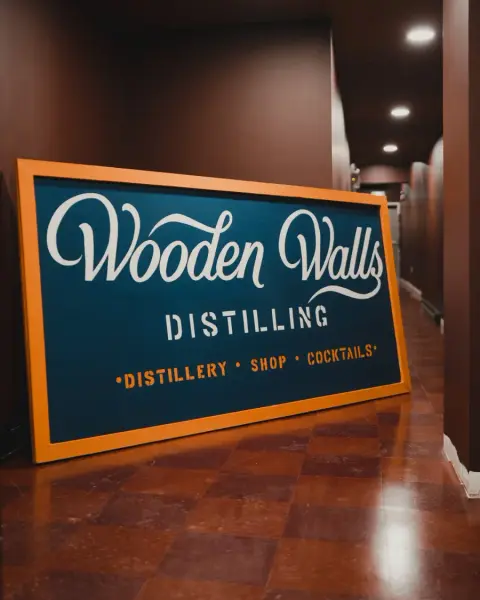 Wooden Walls Distilling sign leaning against a wall indoors, featuring text for distillery, shop, and cocktails, with brown walls and tiled 